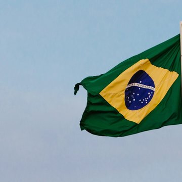 Die Flagge Brasiliens hängt an einer Fahnenstange. Im Hintergrund ist der Himmel zu sehen. Die Flagge hat eine grüne Grundfläche in der ein gelbes Quadrat zu sehen ist. In diesem gelben Quadrat ist eine blaue Kugel zu sehen, in der die Aufschrift "Ordem e Progresso" steht.