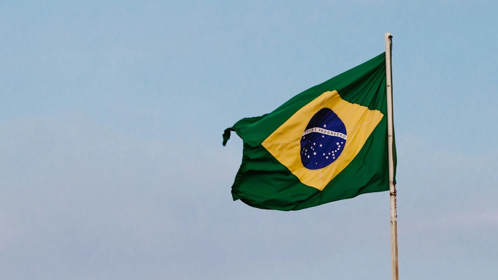 Die Flagge Brasiliens hängt an einer Fahnenstange. Im Hintergrund ist der Himmel zu sehen. Die Flagge hat eine grüne Grundfläche in der ein gelbes Quadrat zu sehen ist. In diesem gelben Quadrat ist eine blaue Kugel zu sehen, in der die Aufschrift "Ordem e Progresso" steht.
