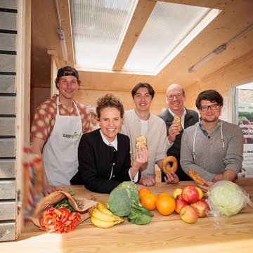 Fünf fröhliche Menschen schauen aus dem Fairteiler. Auf einer Theke liegt Obst und Gemüse.