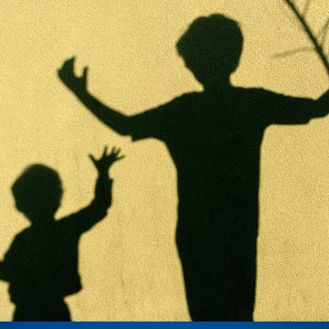 eine gelbe Mauer auf der der Schatten eines größeren und kleineren Kindes zu sehen sind, die beide ihre Arme nach oben heben.