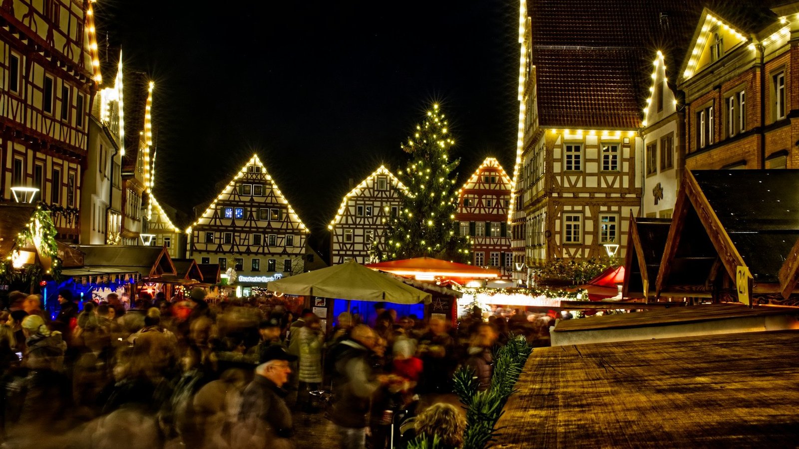 Häuser an denen Lichterketten angebracht sind. Viele Menschen laufen zwischen aufgebauten Ständen durch. 