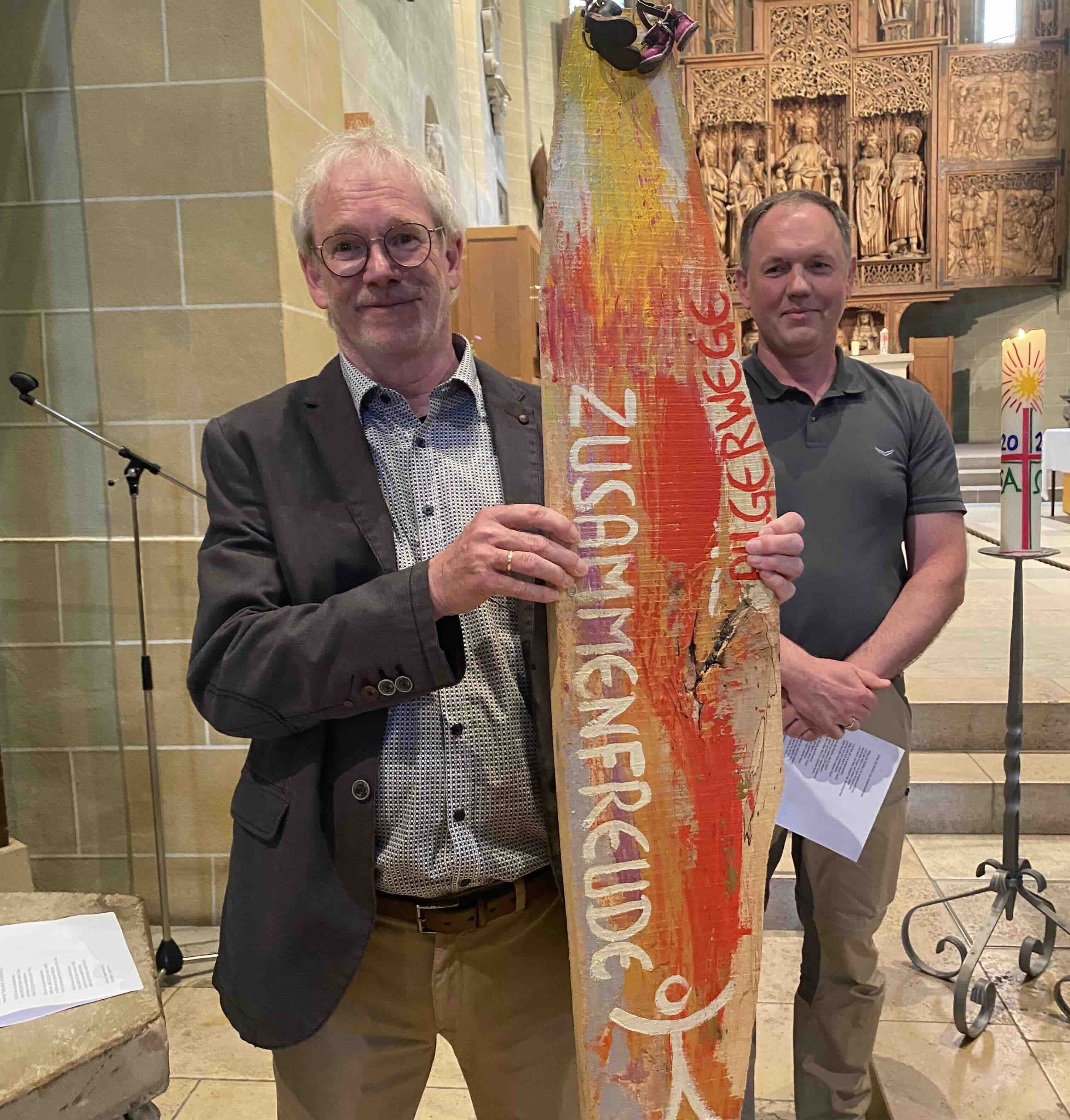 Jürgen Rist steht in der Stadtkirche Winnenden und hält eine Wegetafel aus Holz in der Hand.