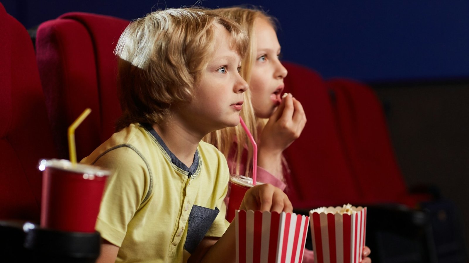 Zwei Kinder sitzen in einem Kinosaal in roten Sesseln. Sie haben Popcorntüten auf dem Schoß