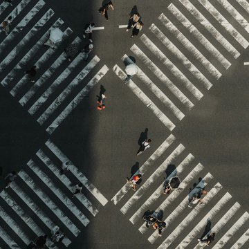 Menschen überqueren eine Straße von vier Seiten über weiße Streifen (Zebrastreifen). Das Foto ist aus der Vogelperspektive von oben aufgenommen.