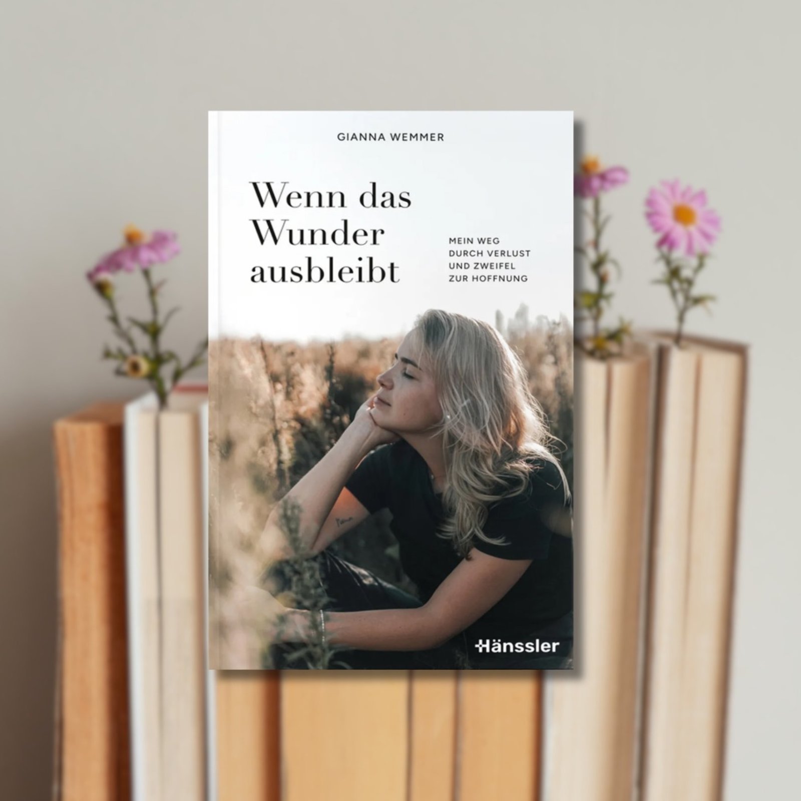 Buchcover "Gianna Wemmer: Wenn das Wunder ausbleibt" im Vordergrund, Bücher, in die rosa Blumen gesteckt sind, im Hintergrund
