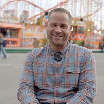Lucas Zehnle sitzt in einem Rollstuhl und lächelt in die Kamera. Im Hintergrund sind Fahrgeschäfte des Cannstatter Wasens in Stuttgart zu sehen.