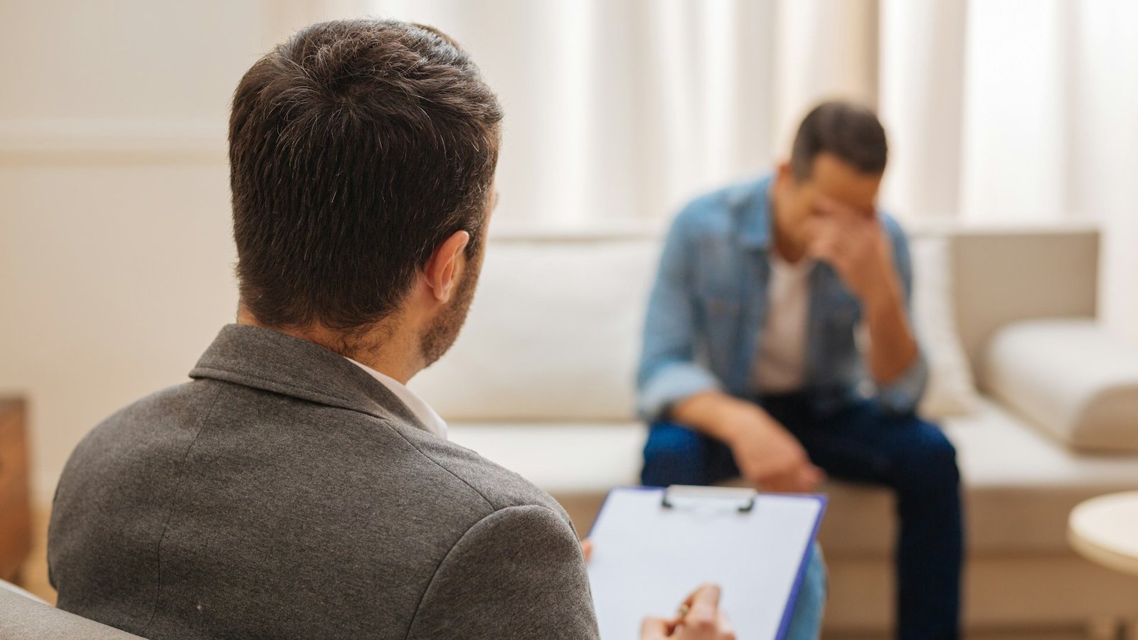 Ein Mann sitz auf einem Sofa und schlägt die Hand vors Gesicht. Ihm gegenüber sitzt sein Berater in einem braunen Blazer. Den Berater sieht man nur von hinten.