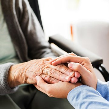 Eine nicht erkennbare Frau sitzt im Rollstuhl. Eine andere jüngere Person hält die Hände der Frau.