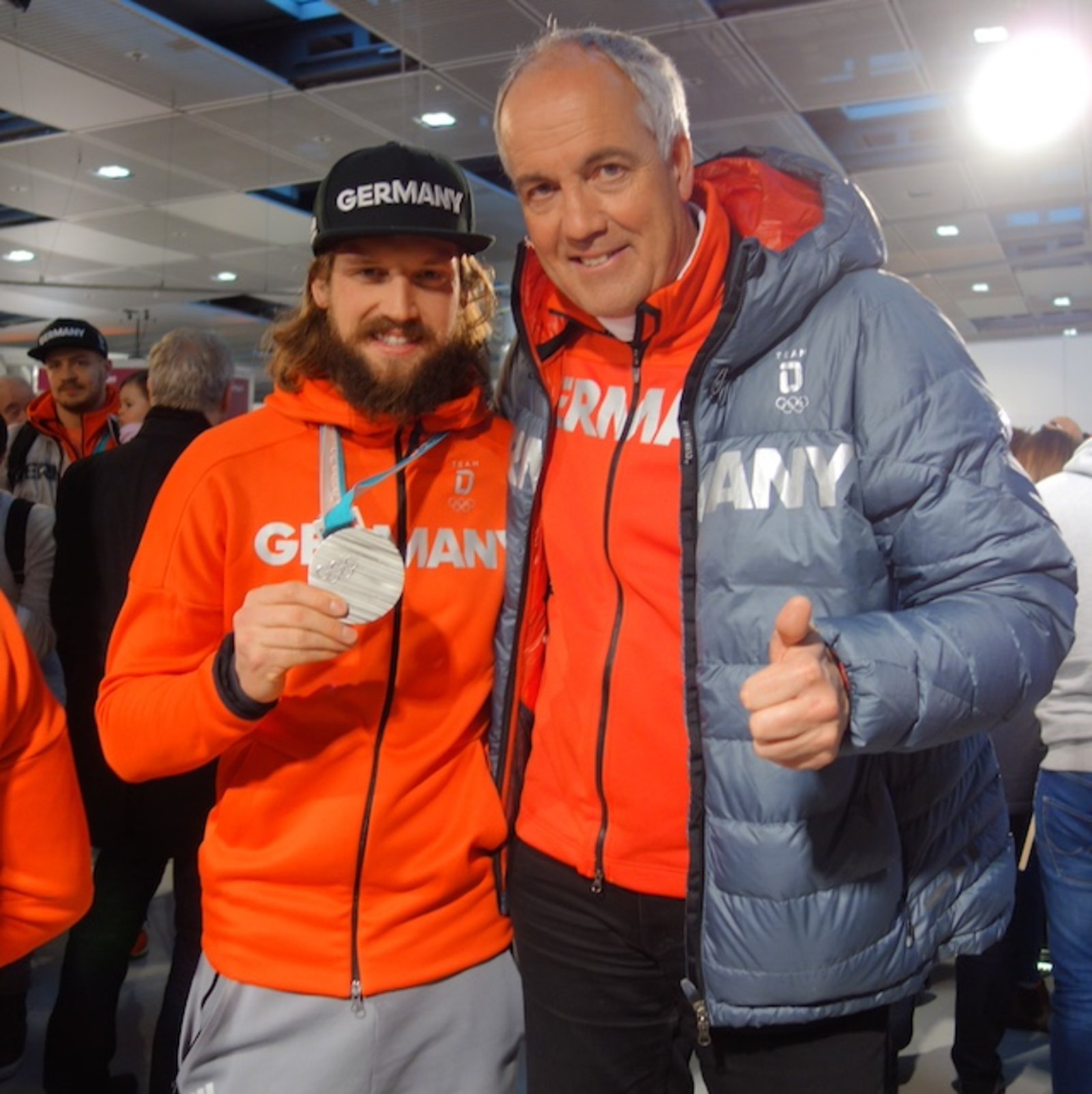 Eishockeyspieler Timo Pielmeier hält seine Silbermedaille in der Hand, neben ihm Olympia-Pfarrer Thomas Weber.