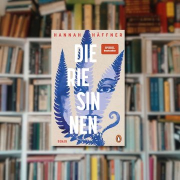 Buchtipp: „Die Riesinnen“ im Vordergrund. Im Hintergrund ist ein gut gefülltes Bücherregal zu sehen.