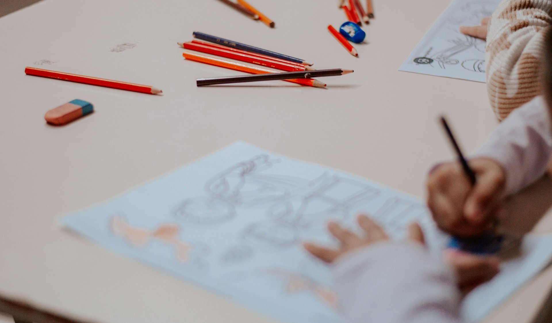 Auf einer hellen Tischplatte liegen rote und orangefarbene Holzbuntstifte. Kinderhände malen auf zwei weißen Papierblättern.