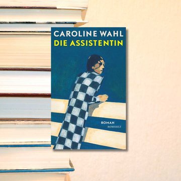 Buchcover „Die Assistentin” von Caroline Wahl. Im Hintergrund ist ein Bücherstapel zu sehen vor einem hellrosa-beigen Hintergrund.