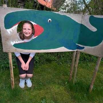 Ein Mädchen schaut aus einem ausgeschnittenen Loch aus einem Walkopf heraus. Der Wal ist auf eine große Holztafel gemalt