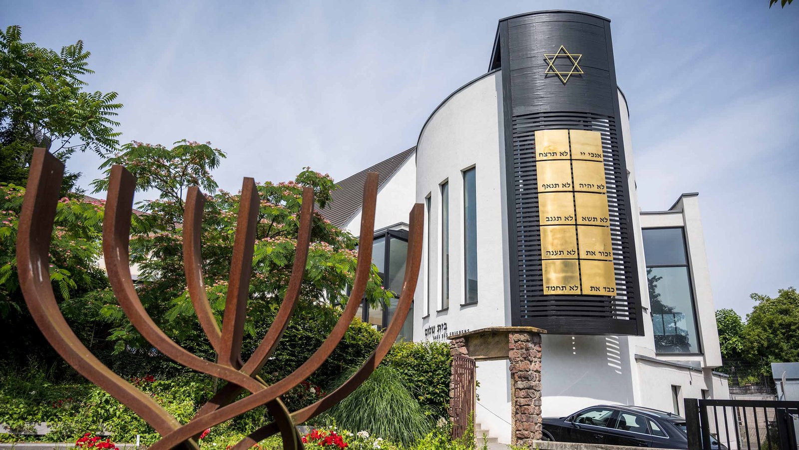 Das Foto zeigt die Synagoge Beith-Shalom der jüdischen Kultusgemeinde von Rheinland-Pfalz in Speyer. Im linken Bildvordergrund ist ein stilisierter Siebenarmiger Leuchter zu sehen. Im rechten Bildhintergrund das Gebäude der Synagoge.
