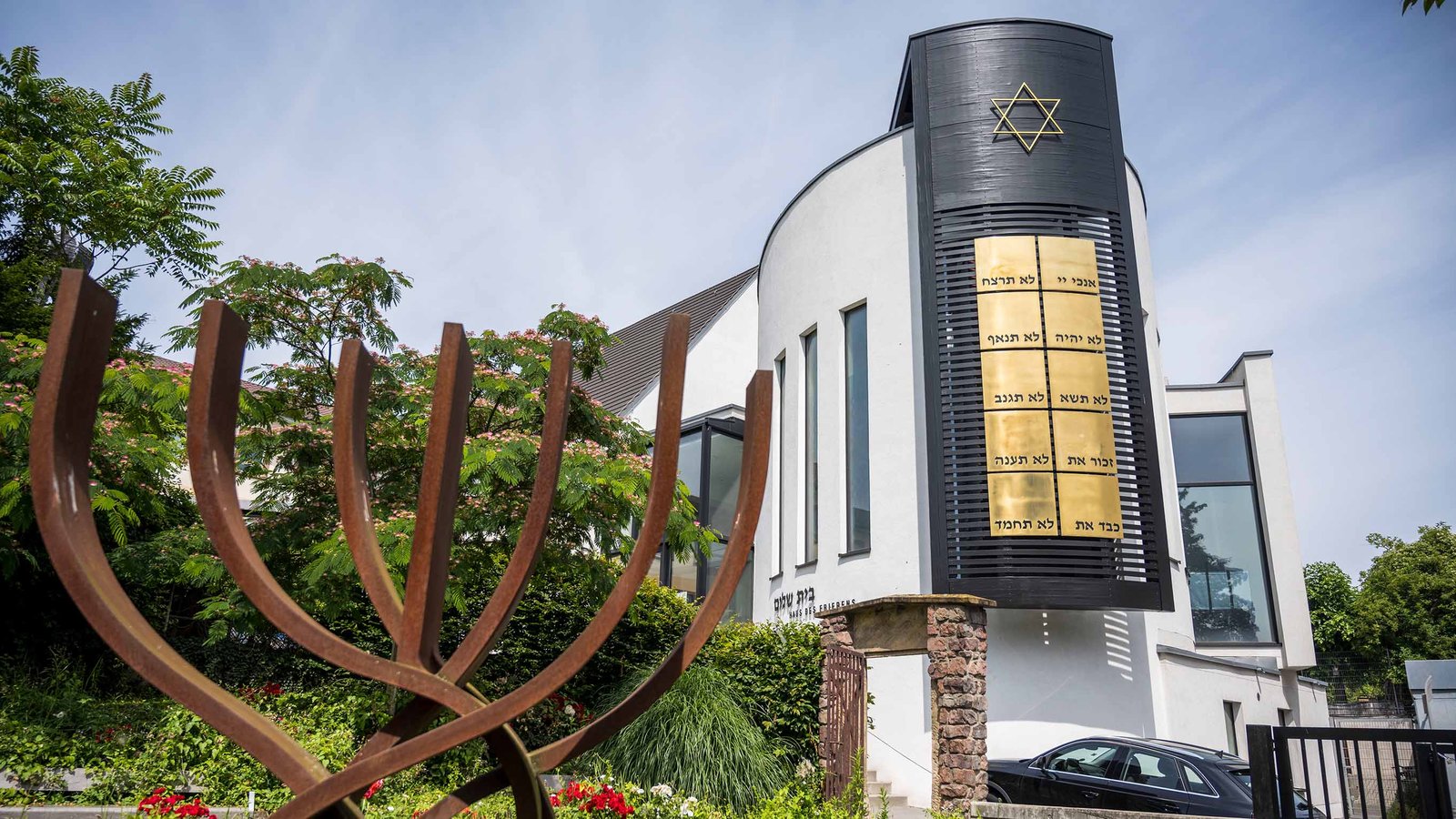 Das Foto zeigt die Synagoge Beith-Shalom der jüdischen Kultusgemeinde von Rheinland-Pfalz in Speyer. Im linken Bildvordergrund ist ein stilisierter Siebenarmiger Leuchter zu sehen. Im rechten Bildhintergrund das Gebäude der Synagoge.