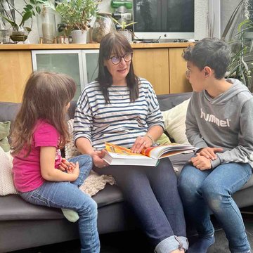 Eine Frau liest zwei Kindern vor. Sie sitzen auf einem Sofa. 