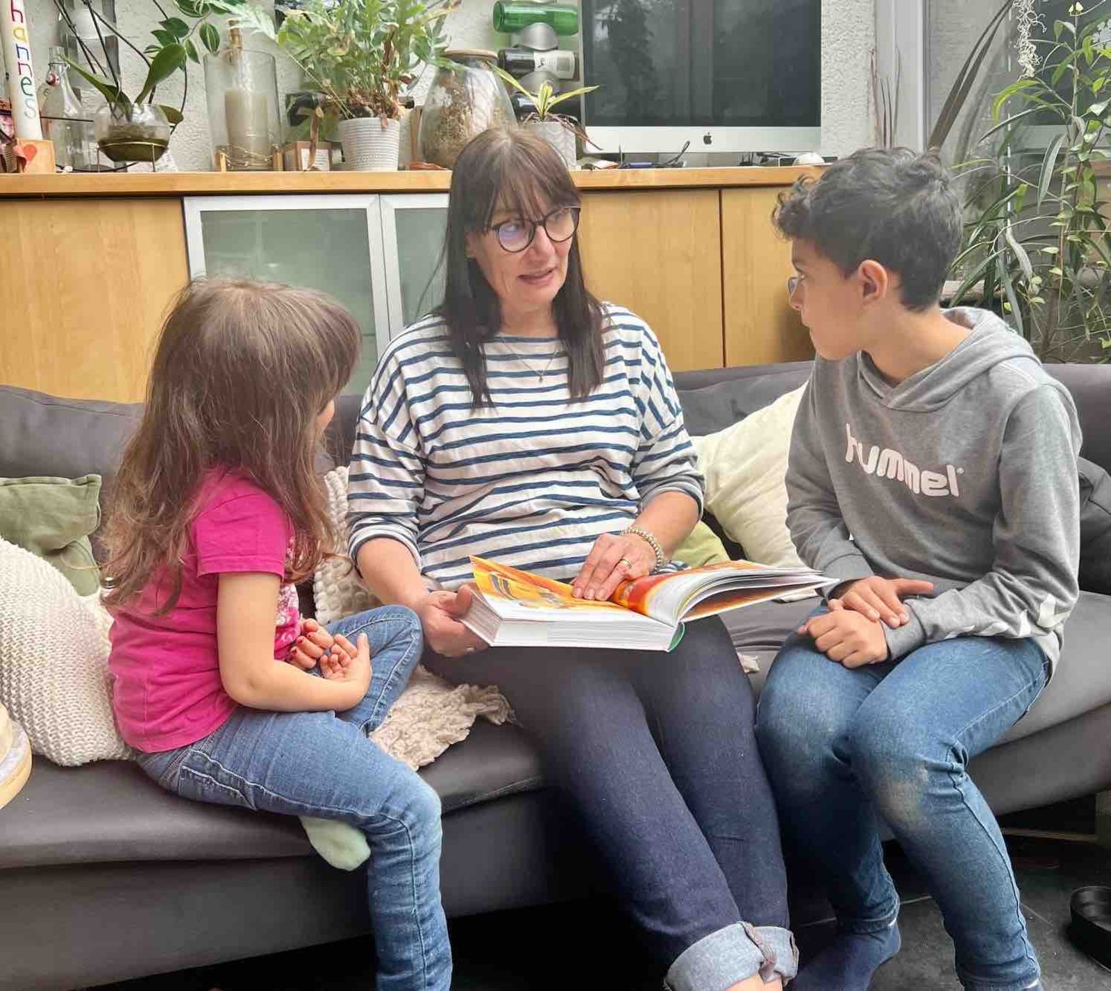 Eine Frau liest zwei Kindern vor. Sie sitzen auf einem Sofa. 
