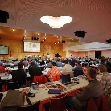 Im Stuttgarter Hospitalhof sitzen die Synodalabgeordneten mit Laptops an den Tischen. Vorne ist eine kleine Bühne mit einem Bildschirm und einem Rednerpult zu sehen.