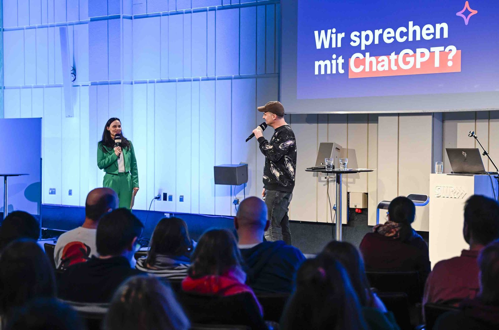 Ein Mann und eine Frau stehen auf einer Bühne. Hinter ihnen steht auf einer Leinwand: "Wir sprechen mit ChatGPT"