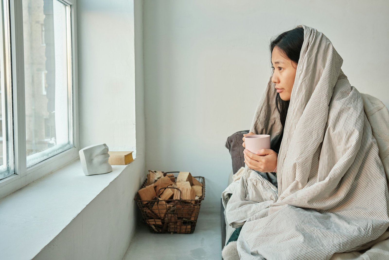 Frau sitzt in eine Decke gehüllt mit einer Tasse in der Hand am Fenster.