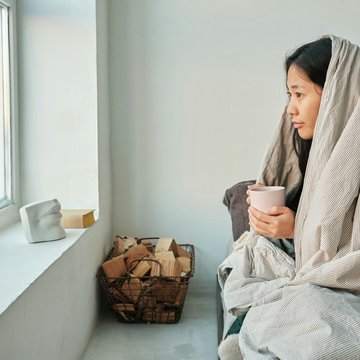 Frau sitzt in eine Decke gehüllt mit einer Tasse in der Hand am Fenster.