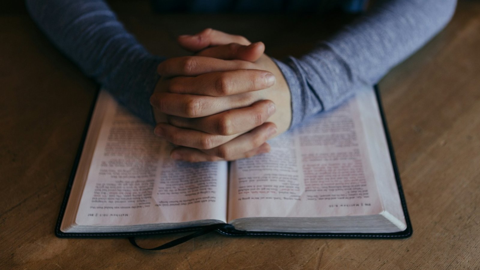 Eine aufgeschlagene Bibel liegt auf einem Tisch. Eine Person sitzt an dem Tisch, hat ihre Hände auf der Bibel wie zum Gebet gefaltet.
