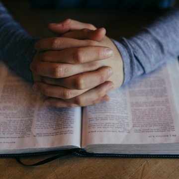 Eine aufgeschlagene Bibel liegt auf einem Tisch. Eine Person sitzt an dem Tisch, hat ihre Hände auf der Bibel wie zum Gebet gefaltet.
