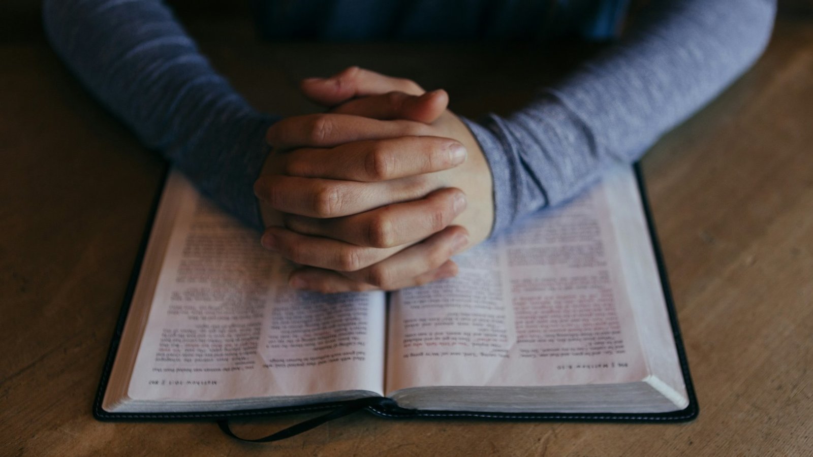 Eine aufgeschlagene Bibel liegt auf einem Tisch. Eine Person sitzt an dem Tisch, hat ihre Hände auf der Bibel wie zum Gebet gefaltet.