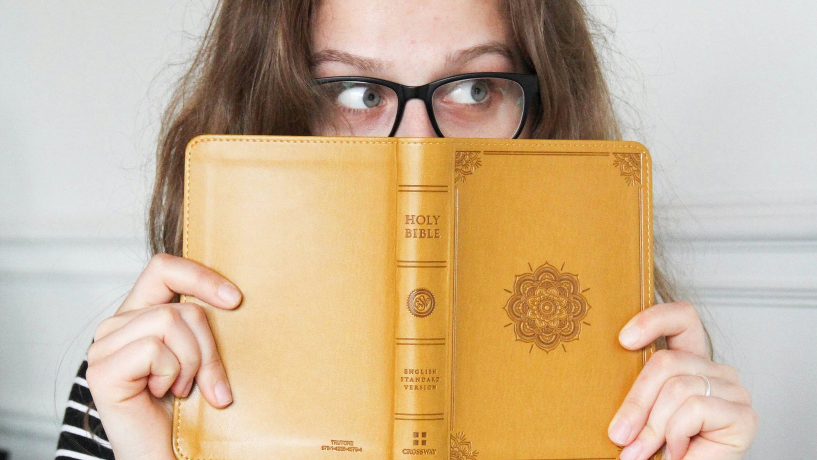 Eine junge Frau hält sich eine geöffnete Bibel mit gelbem Umschlag vor ihr Gesicht. Sie trägt eine Brille und blickt hinter der Bibel hervor. Ihr Blick geht nach rechts.
