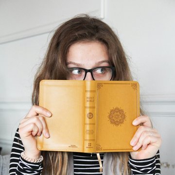 Eine junge Frau hält sich eine geöffnete Bibel mit gelbem Umschlag vor ihr Gesicht. Sie trägt eine Brille und blickt hinter der Bibel hervor. Ihr Blick geht nach rechts.