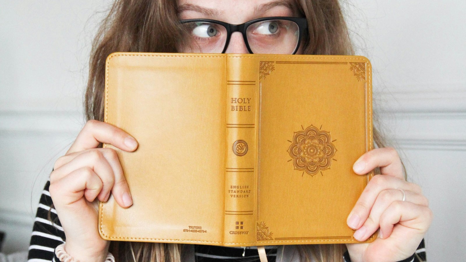 Eine junge Frau hält sich eine geöffnete Bibel mit gelbem Umschlag vor ihr Gesicht. Sie trägt eine Brille und blickt hinter der Bibel hervor. Ihr Blick geht nach rechts.
