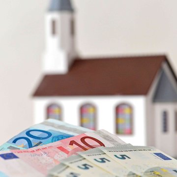 Symbolbild: Im Hintergrund ist das Modell einer kleinen Kirche zu sehen, im Vordergrund liegen Geldscheine. 