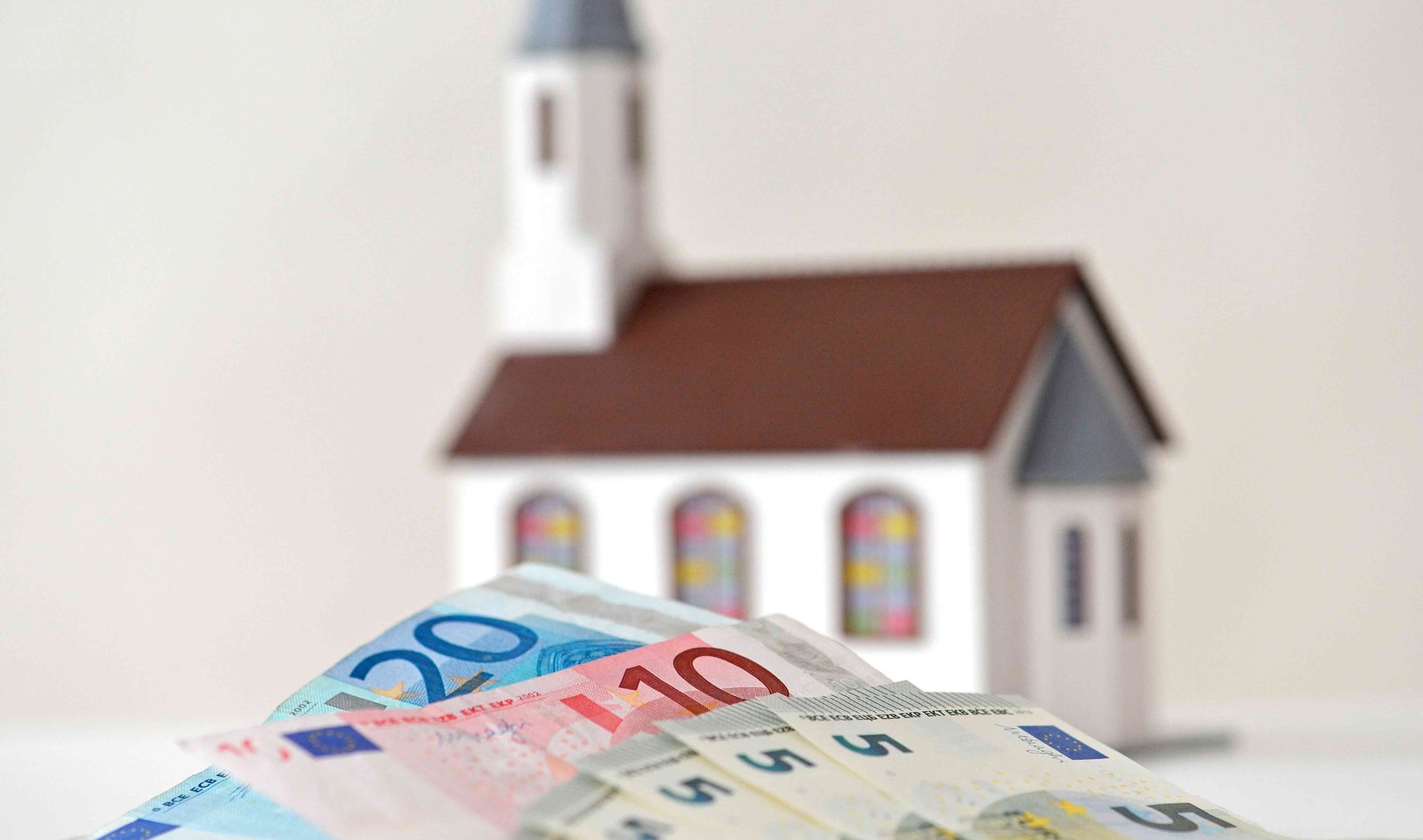 Symbolbild: Im Hintergrund ist das Modell einer kleinen Kirche zu sehen, im Vordergrund liegen Geldscheine. 