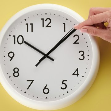 Eine analoge runde Uhr vor einem gelben Hintergrund. Eine Frauenhand zeigt auf den Minutenzeiger.