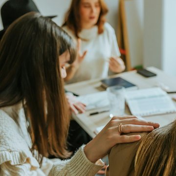 Eine Gruppe von Frauen sitzt gemeinsam an einem Tisch, eine Frau senkt ihren Kopf und hat eine Hand auf die Schulter einer anderen Frau gelegt.