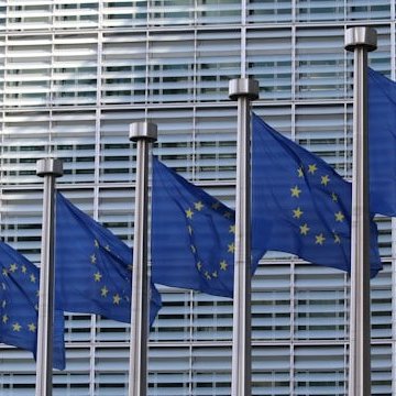 Vor einem Gebäude sind nebeneinander sieben Europaflaggen (Blauer Hintergrund mit gelben Sternen, die kreisförmig angeordnet sind) an Fahnenstangen angebracht.