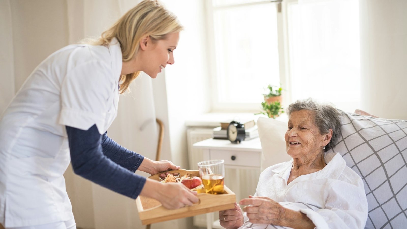 Pflegekraft bringt Seniorin Frühstück ans Bett.