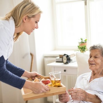 Pflegekraft bringt Seniorin Frühstück ans Bett.