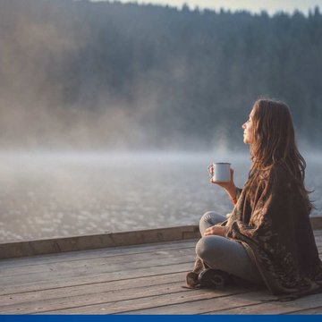 Eine Frau sitzt an einem Steg im Wald. Sie hält eine Tasse in der Hand und blickt über einen nebelverhangenen See.