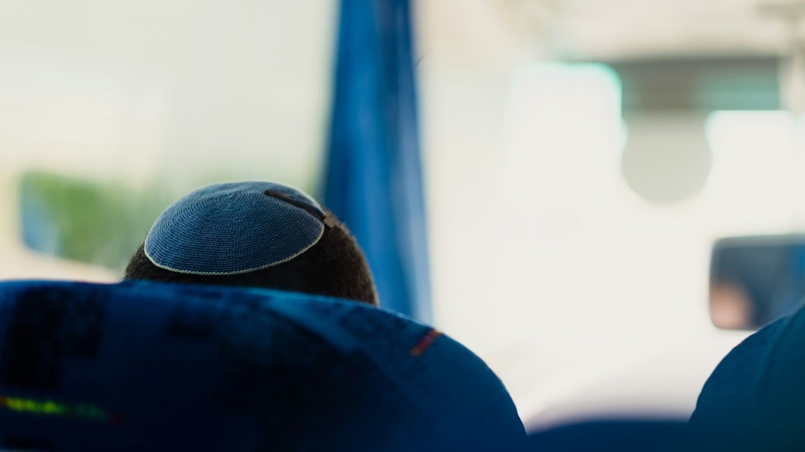 Das Bild zeigt den Hinterkopf eines Mannes, der in einem Bus sitzt und auf dessen Kopf eine Kippa (jüdische Kopfbedeckung für Männer) mit einer Haarspange befestigt ist.