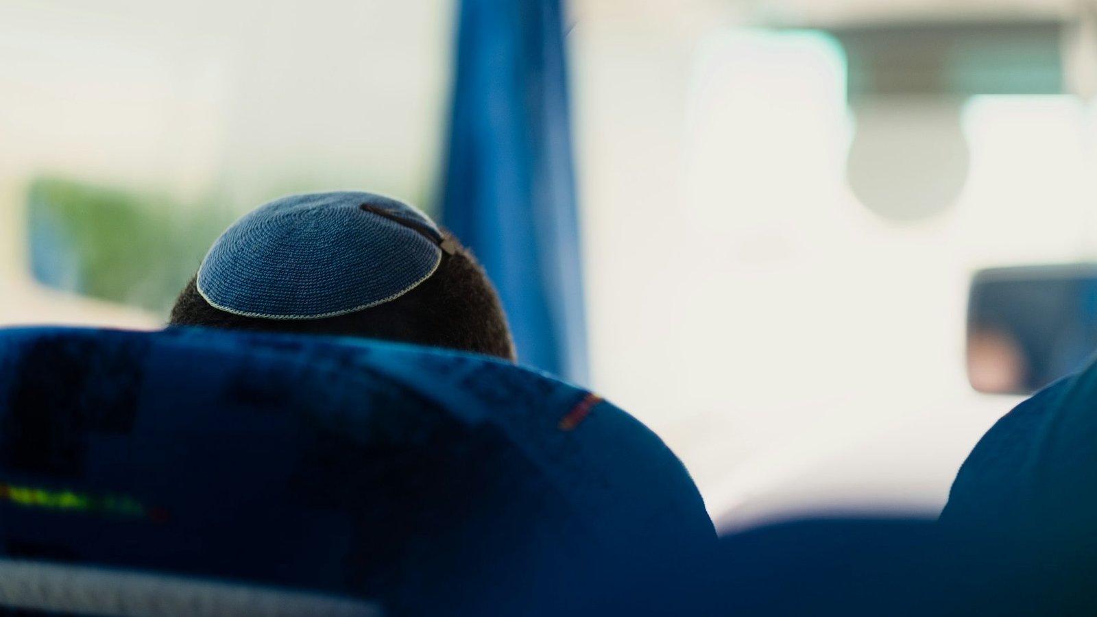 Das Bild zeigt den Hinterkopf eines Mannes, der in einem Bus sitzt und auf dessen Kopf eine Kippa (jüdische Kopfbedeckung für Männer) mit einer Haarspange befestigt ist.