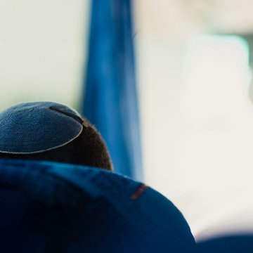 Das Bild zeigt den Hinterkopf eines Mannes, der in einem Bus sitzt und auf dessen Kopf eine Kippa (jüdische Kopfbedeckung für Männer) mit einer Haarspange befestigt ist.