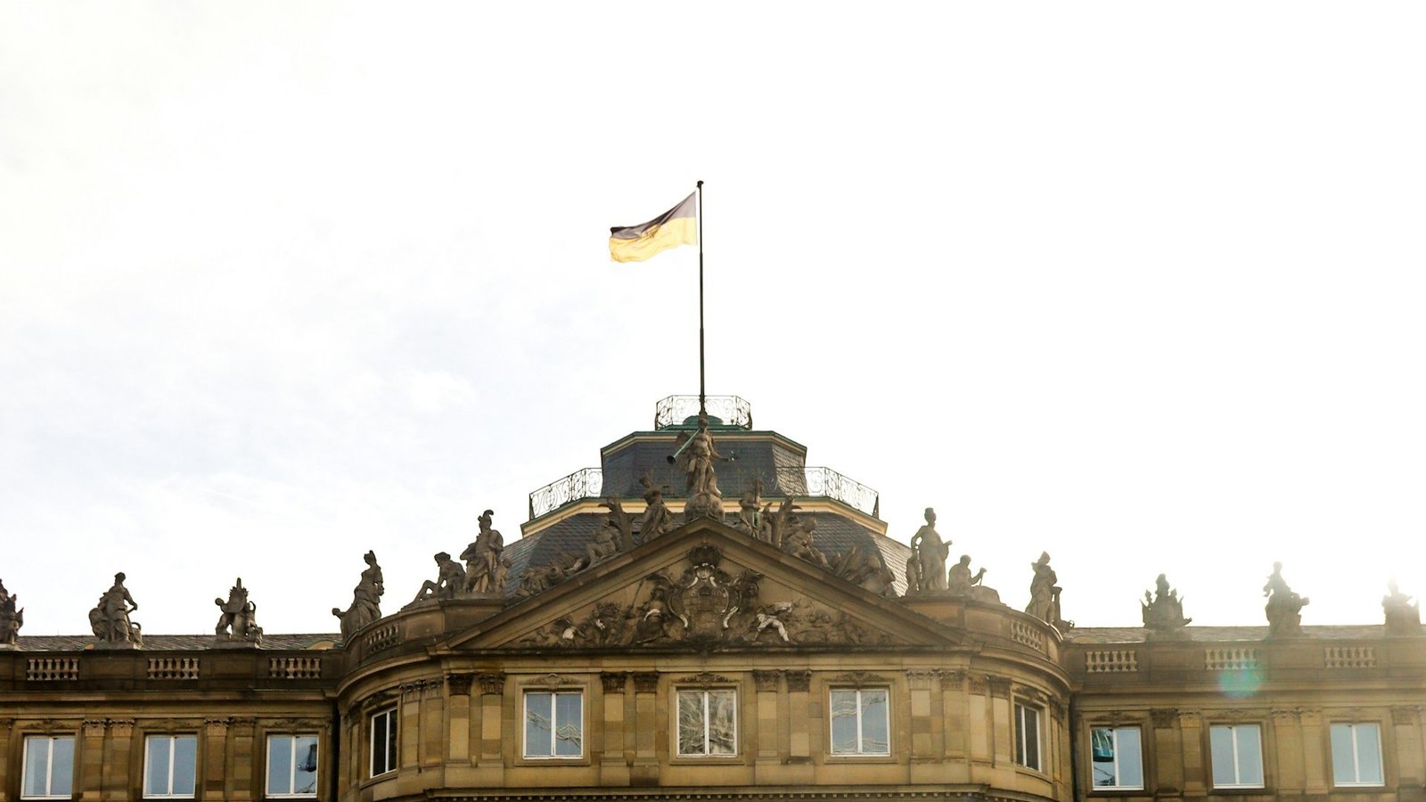 Das Foto zeigt das Schloss von Stuttgart. Davor ist ein Springbrunnen zu sehen. Auf dem Gebäude ist die schwarz-gelbe Flagge von Baden-Württemberg gehisst.