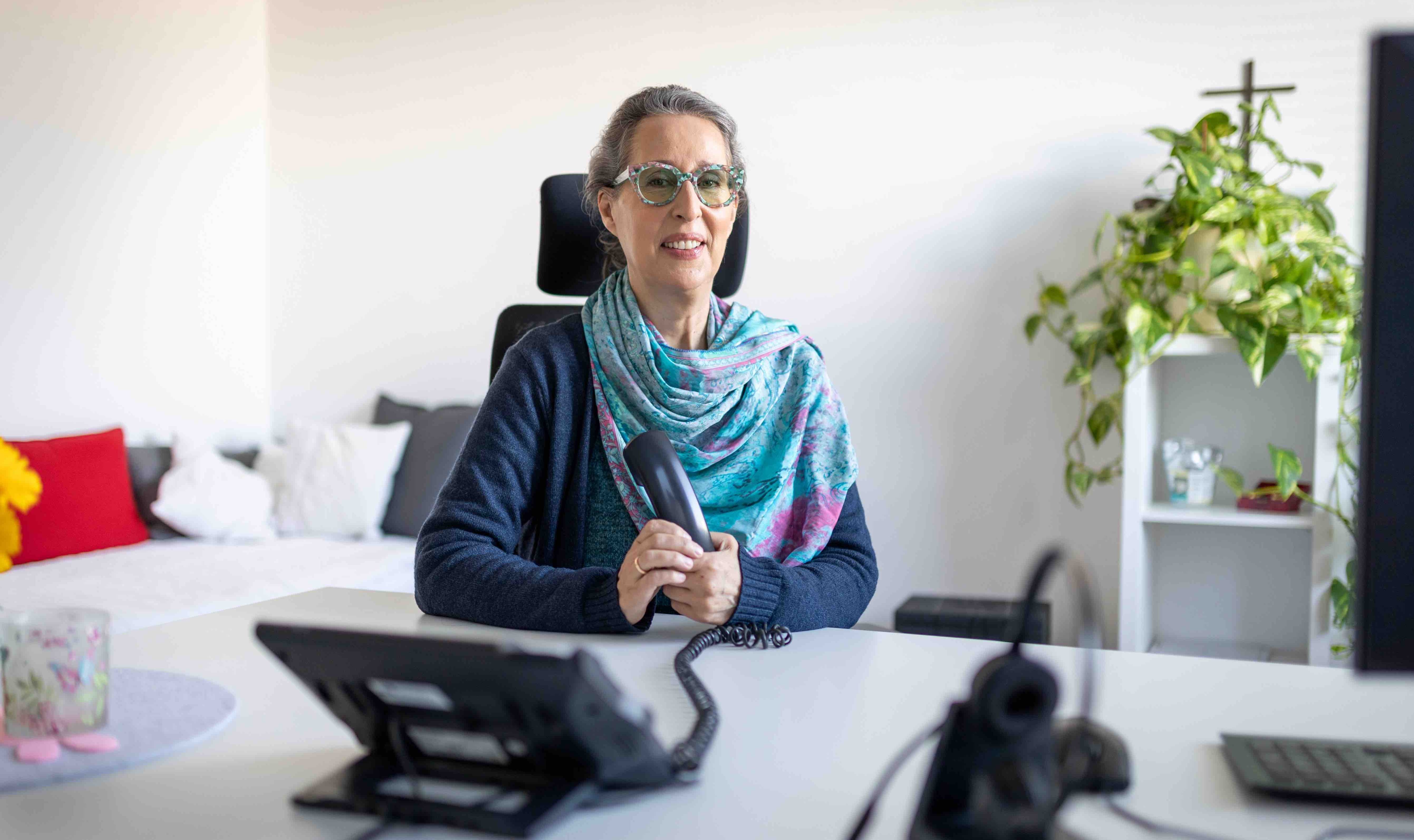 Eine Frau (Beate) sitzt an einem Schreibtisch, sie hält einen Telefonhörer in der Hand und lächelt.