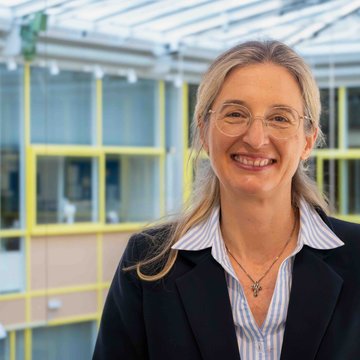 Das Foto zeigt Nina Lubomierski. Sie lächelt in die Kamera, trägt eine Bluse und einen schwarzen Blazer darüber. Sie hat lange blonde Haare und trägt eine Brille.