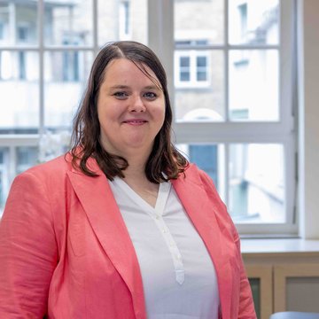 Lea Blattner trägt eine weiße Bluse, ein pinkes Jackett und lächelt in die Kamera. Im Hintergrund sind Fenster zu sehen und zwei Stühle.