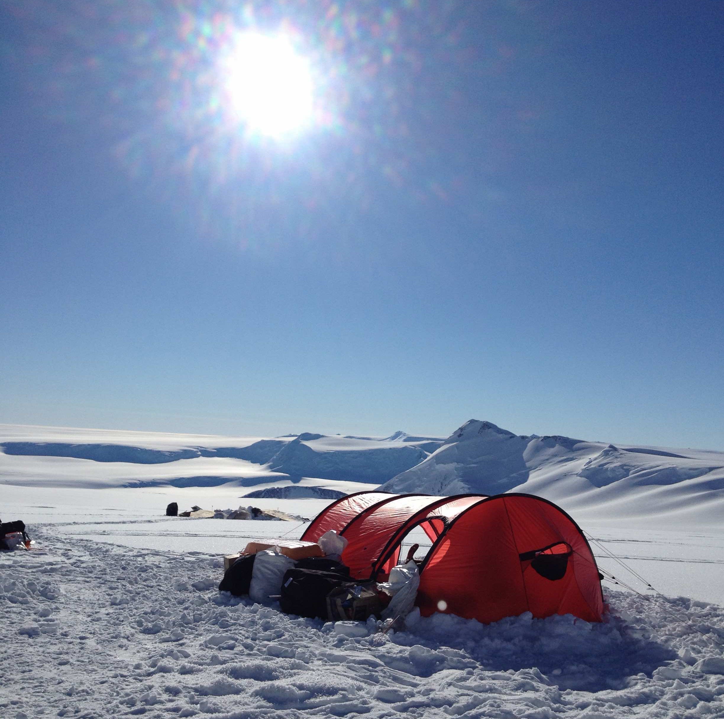 Ein rotes Zelt steht aufgebaut in einem Schneefeld. Im blauen Himmel ist die Sonne zu sehen.