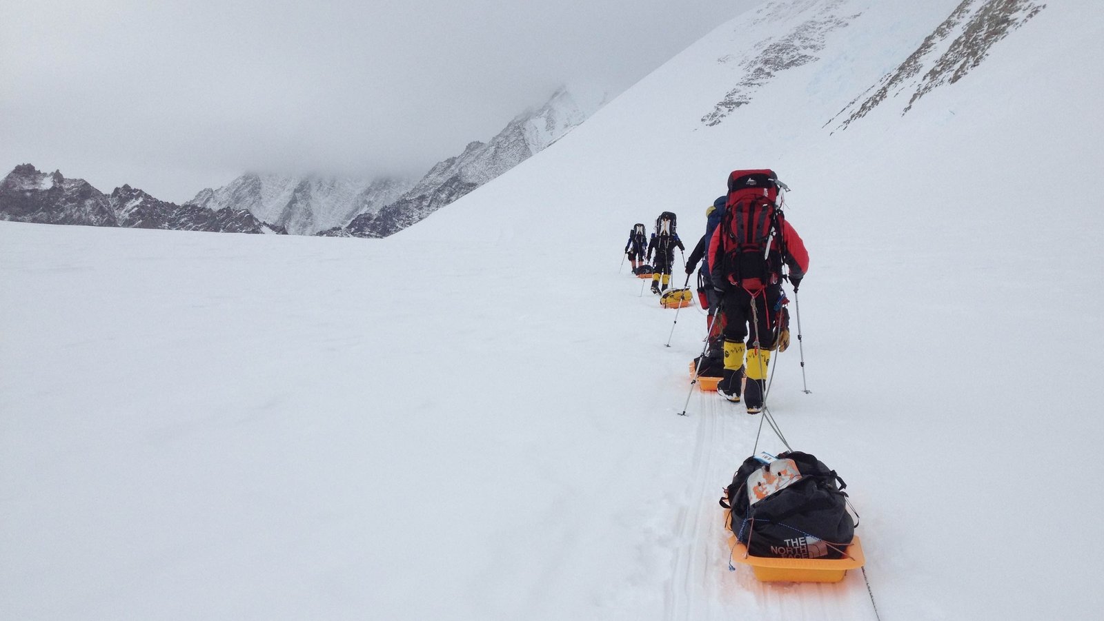 Eine Karawane von Menschen läuft in Ausrüstung durch ein Schneefeld, rechts sind schneebedeckte Berge zu sehen. Der letzte Mensch in der Karawane hat einen großen Rucksack auf und zieh eine beladene Wanne hinter sich her.