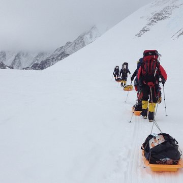 Eine Karawane von Menschen läuft in Ausrüstung durch ein Schneefeld, rechts sind schneebedeckte Berge zu sehen. Der letzte Mensch in der Karawane hat einen großen Rucksack auf und zieh eine beladene Wanne hinter sich her.