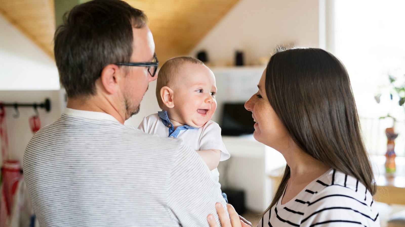 Eine junge Familie zu Hause. Der Vater hält ein lachendes Baby auf dem Arm. Die Mutter und das Baby schauen sich an. 